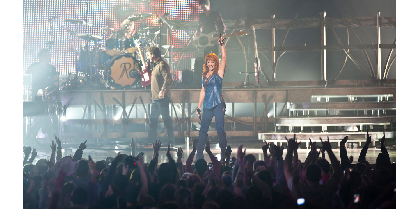 Country music singer Reba McEntire on stage singing a song with the crowd in front of her with her hand up, dancing.